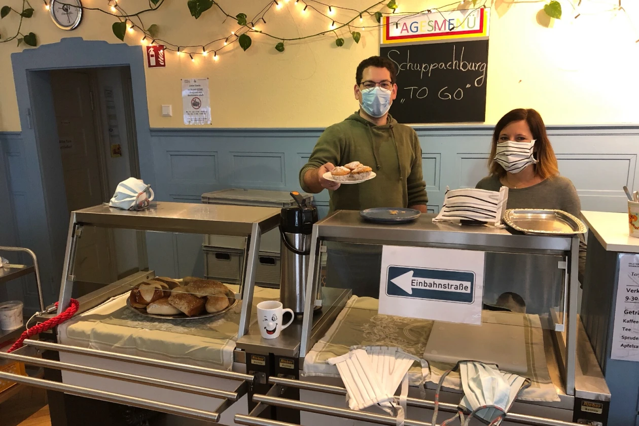 Zwei Personen stehen hinter einer Theke mit Glasaufsatz. Beide tragen Mund-Nasenschutz. Der Mann rechts h&auml;lt einen Teller mit Muffins in der Hand. Im Hintergrund h&auml;ngt eine Tafel mit der Aufschrift "Schuppachburg TO GO".