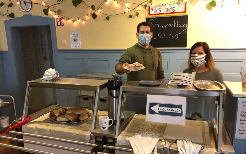Zwei Personen stehen hinter einer Theke mit Glasaufsatz. Beide tragen Mund-Nasenschutz. Der Mann rechts h&auml;lt einen Teller mit Muffins in der Hand. Im Hintergrund h&auml;ngt eine Tafel mit der Aufschrift "Schuppachburg TO GO".