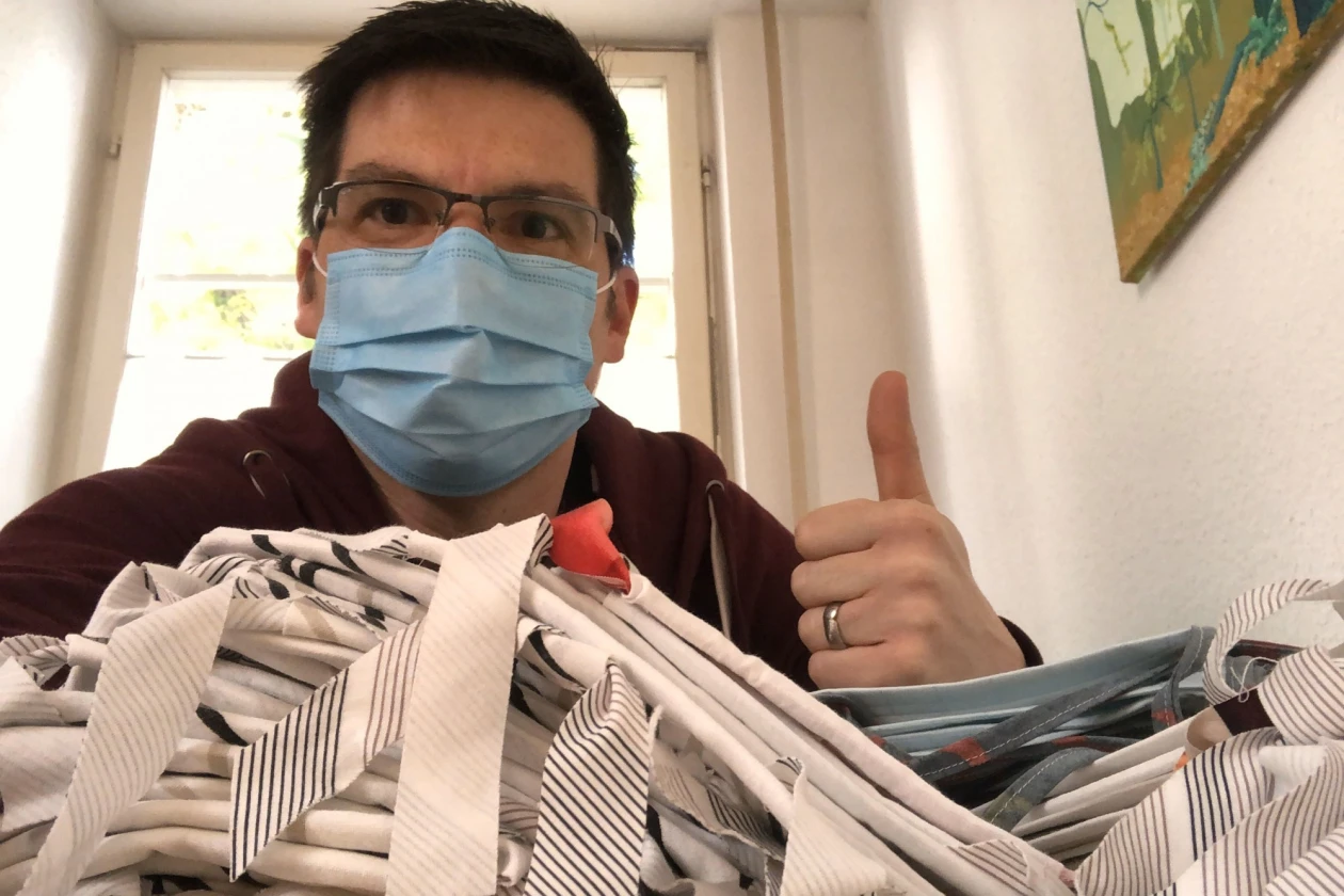 Ein Mann mit Mund-Nasenschutz blickt in die Kamera und hebt den Daumen nach oben. In der anderen Hand h&auml;lt er textile Werkst&uuml;cke.