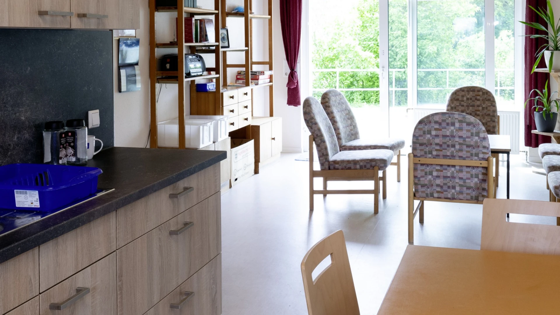 Blick in einen Ess- und Wohnzimmer mit K&uuml;chenzeile. Gro&szlig;e Fenstern und Regale mit B&uuml;chern, sowie eine Pflanze stehen im Hintergrund. und roten Vorh&auml;ngen.