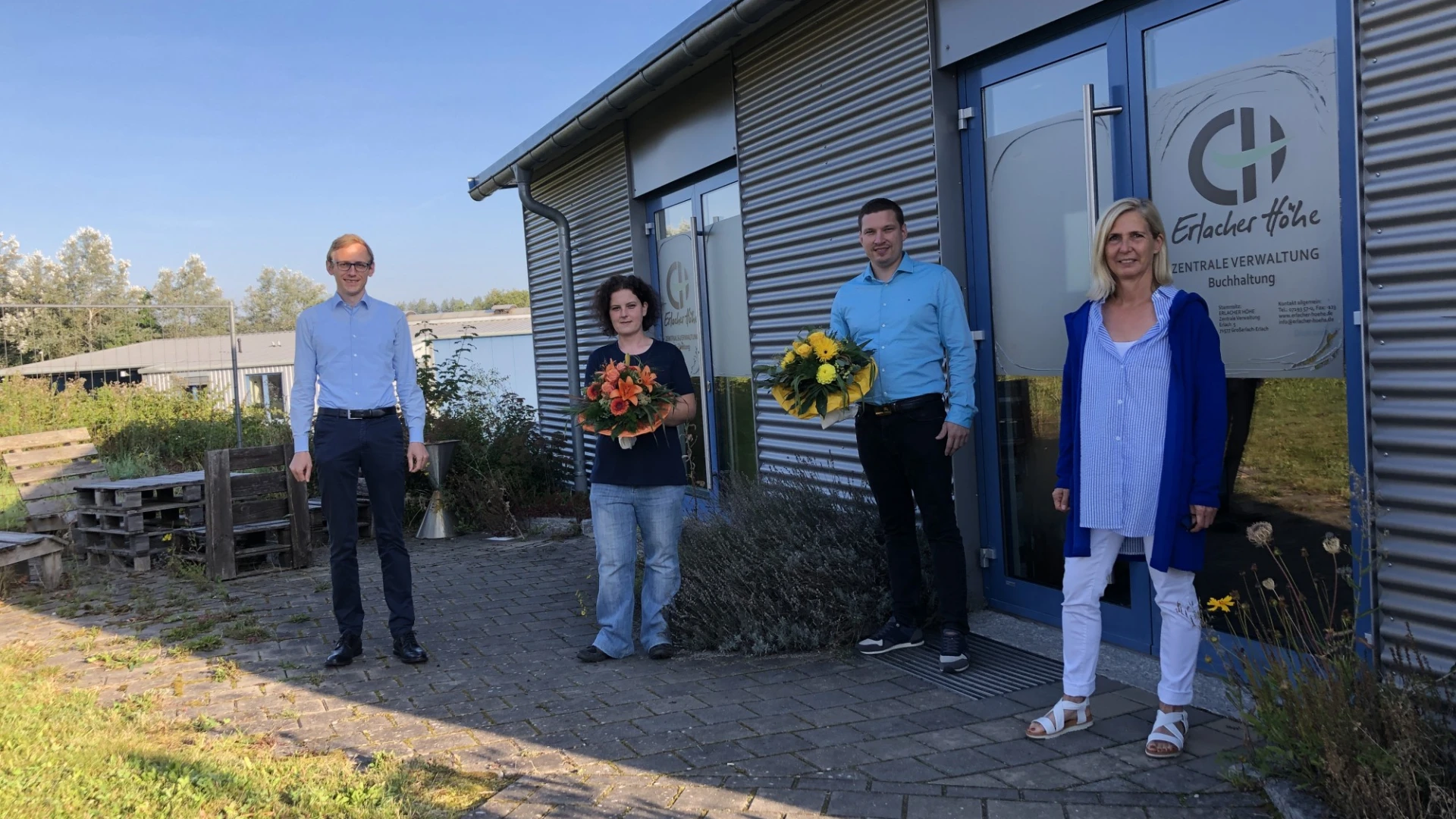 Vier Personen stehen auf dem gepflasterten Eingangsbereich vor einem Geb&auml;ude. Die Frau und der Mann in der Mitte halten einen Blumenstrau&szlig; in der Hand. Auf der Eingangst&uuml;r ist das Logo der Erlacher H&ouml;he zu sehen.