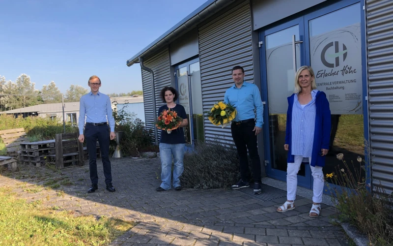Vier Personen stehen auf dem gepflasterten Eingangsbereich vor einem Geb&auml;ude. Die Frau und der Mann in der Mitte halten einen Blumenstrau&szlig; in der Hand. Auf der Eingangst&uuml;r ist das Logo der Erlacher H&ouml;he zu sehen.