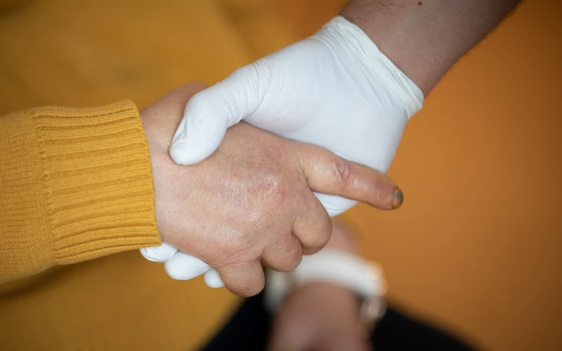 Eine Hand in wei&szlig;em Handschuh und eine Hand einer Person in gelbem Pullover liegen ineinander wie bei einer Begr&uuml;&szlig;ung.