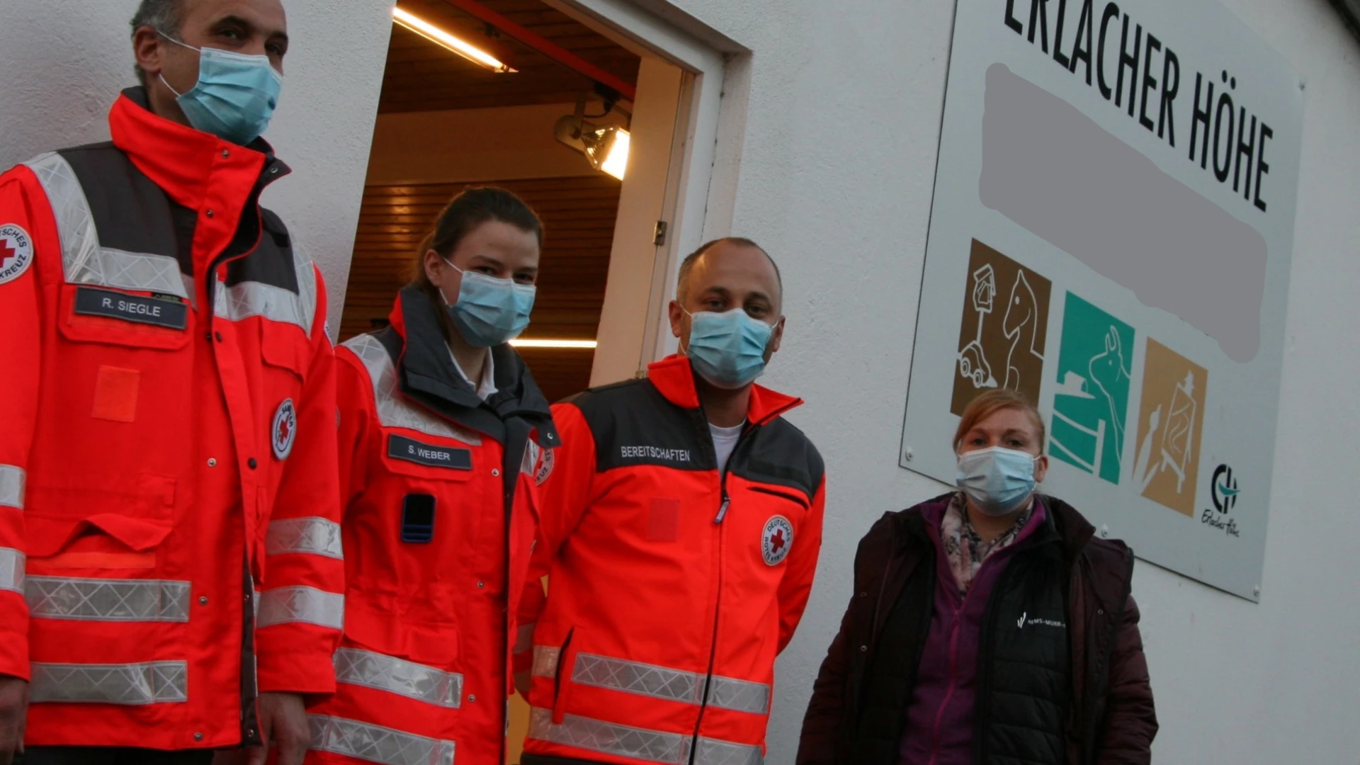 Vier Personen stehen vor einem Geb&auml;ude mit  einem Schild der Erlacher H&ouml;he. Drei von ihnen tragen rote Einsatzjacken des Deutschen Roten Kreuzes und alle tragen Mund-Nasenschutz.