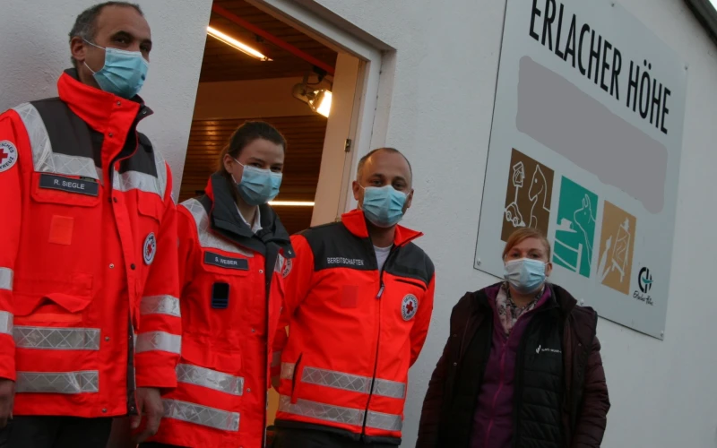 Vier Personen stehen vor einem Geb&auml;ude mit  einem Schild der Erlacher H&ouml;he. Drei von ihnen tragen rote Einsatzjacken des Deutschen Roten Kreuzes und alle tragen Mund-Nasenschutz.