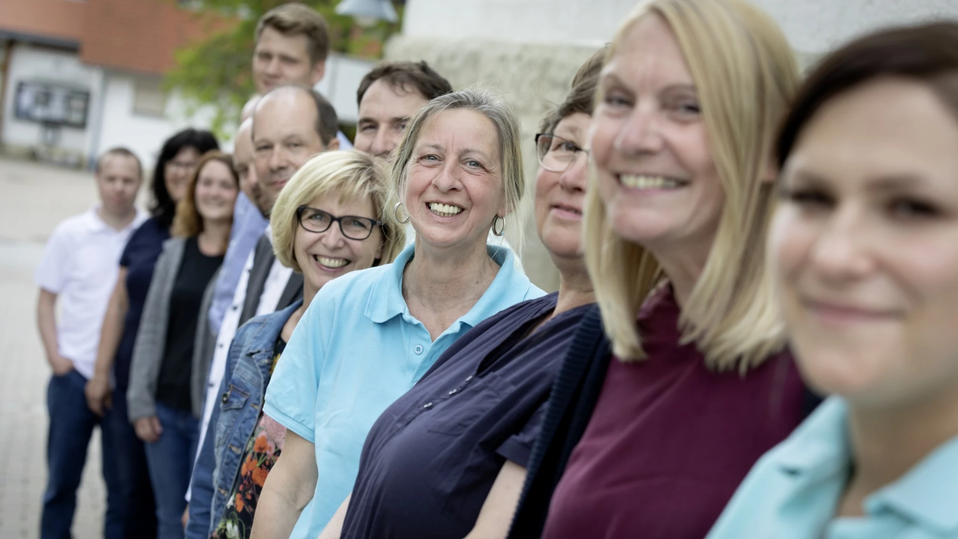 Gruppe von Menschen, die nebeneinander in verschiedenfarbiger Kleidung stehen und fr&ouml;hlich in die Kamera l&auml;cheln. Unscharfer Hintergrund, der ein Geb&auml;ude und eine Mauer zeigt.