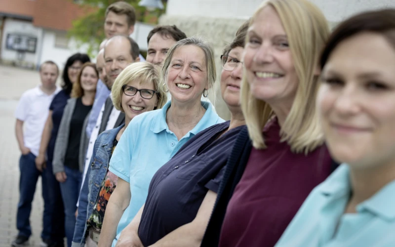 Gruppe von Menschen, die nebeneinander in verschiedenfarbiger Kleidung stehen und fr&ouml;hlich in die Kamera l&auml;cheln. Unscharfer Hintergrund, der ein Geb&auml;ude und eine Mauer zeigt.