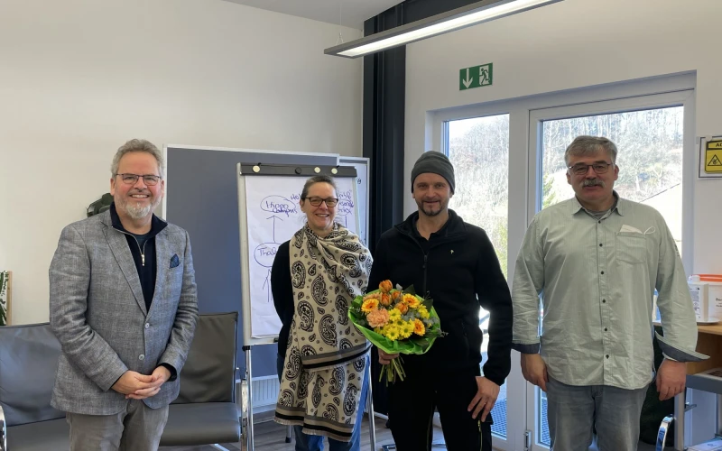 Vier Personen stehen in einem Raum und blicken l&auml;chelnd in die Kamera. Der Mann in der Mitte h&auml;lt einen Blumenstrau&szlig; in der Hand. Im Hintergrund ist eine Pinnwand und St&uuml;hle zu sehen.