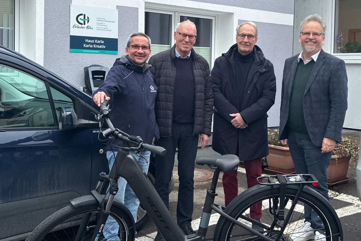 Im Vordergrund des Bildes ist ein neues E-Bike zu sehen. Es ist schwarz und steht auf dem Parkplatz vor einem Geb&auml;ude mit dem Schild Haus Karla. Hinter dem E-Bike stehen vier M&auml;nner und l&auml;cheln in die Kamera.