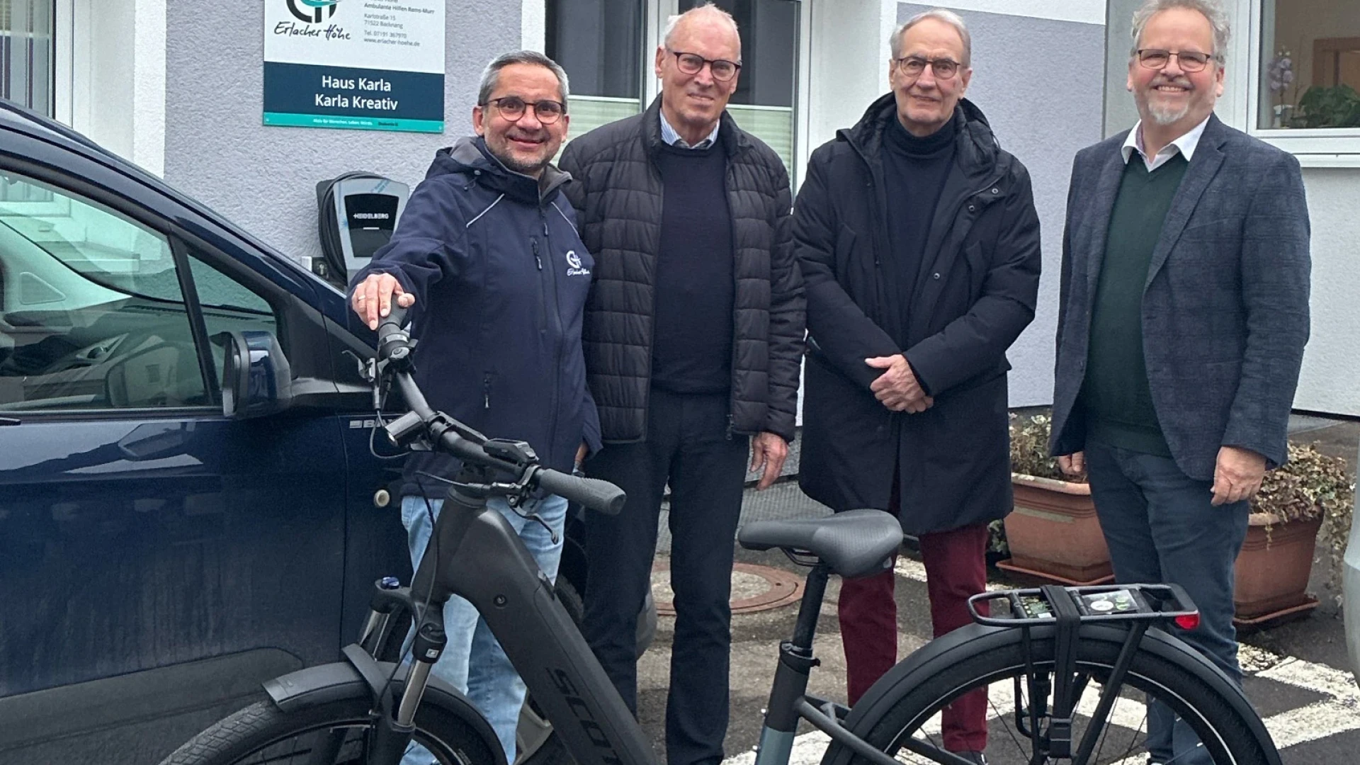 Im Vordergrund des Bildes ist ein neues E-Bike zu sehen. Es ist schwarz und steht auf dem Parkplatz vor einem Geb&auml;ude mit dem Schild Haus Karla. Hinter dem E-Bike stehen vier M&auml;nner und l&auml;cheln in die Kamera.