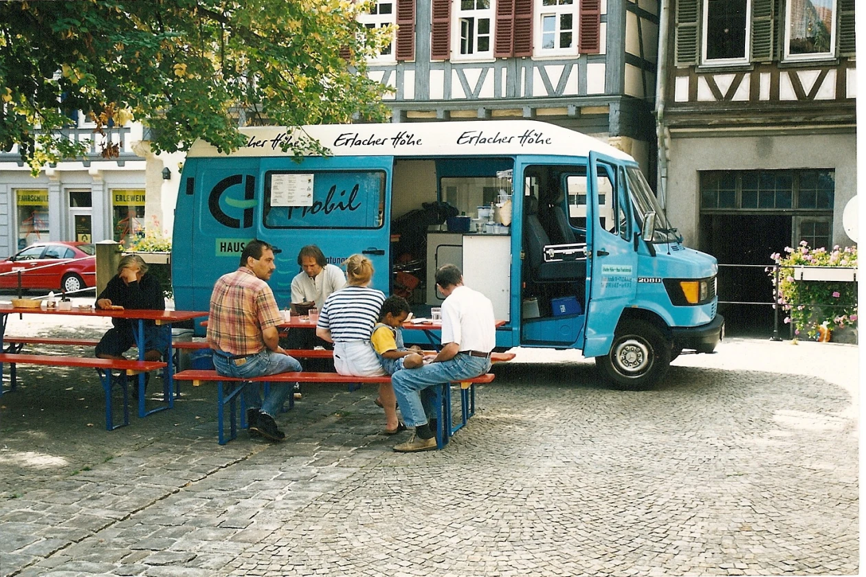 Biertischb&auml;nke mit Menschen vor einem hellblauen Lieferwagen mit der Aufschrift "EH mobil".