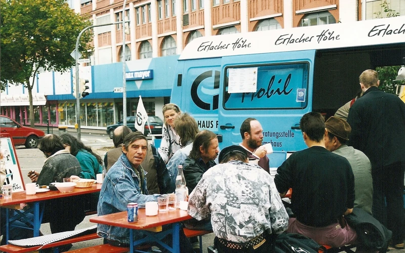 Historische Aufnahme: Menschen an Biertischb&auml;nken mit Getr&auml;nken in einer Stadt vor einem hellblauen Lieferwagen mit der Aufschrift "EH mobil".