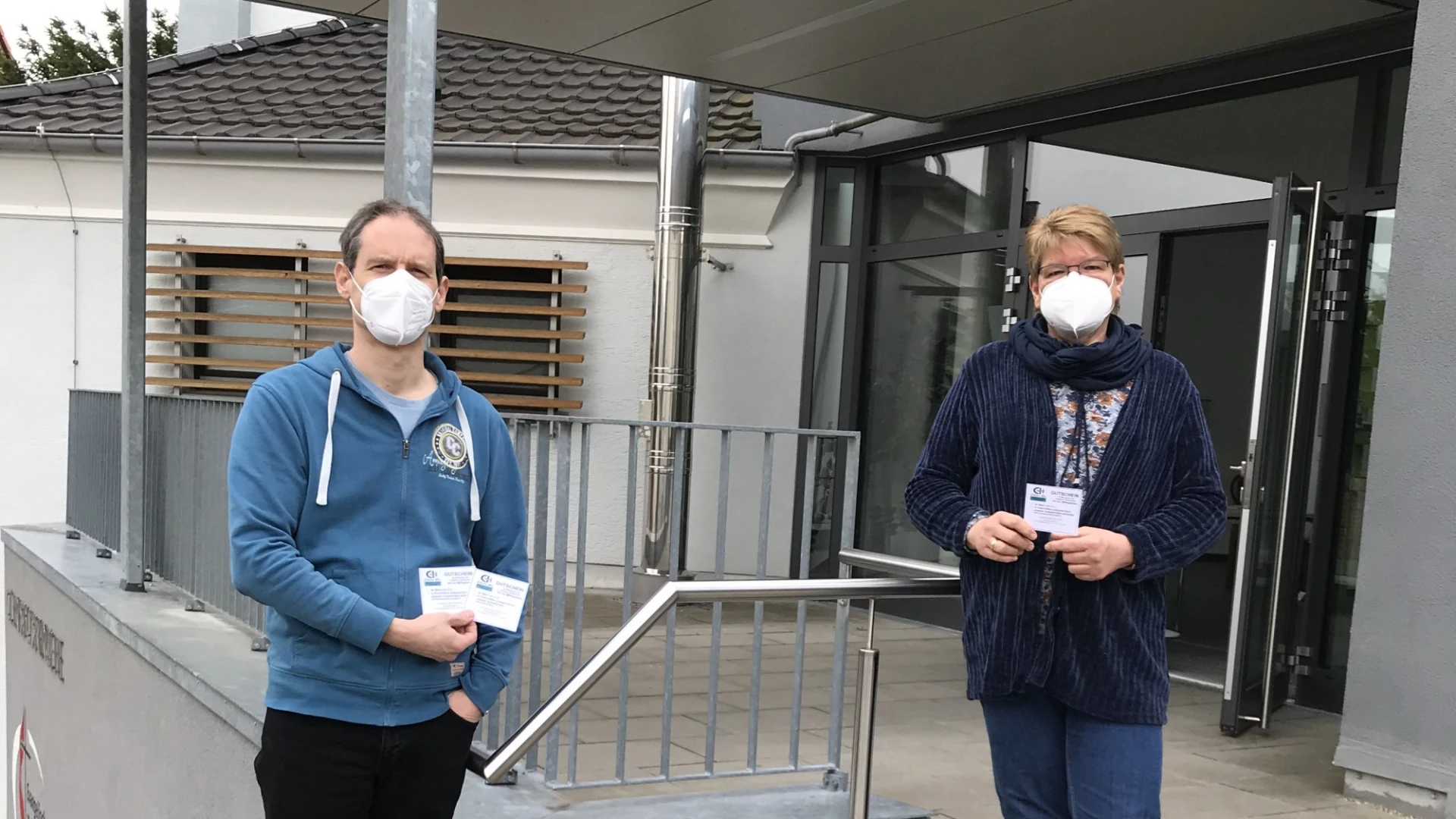 Zwei Personen stehen im Eingangsbereich eines Geb&auml;udes an einem Gel&auml;nder. Sie zeigen kleine Zettel in die Kamera. Beide tragen Mund-Nasenschutz.