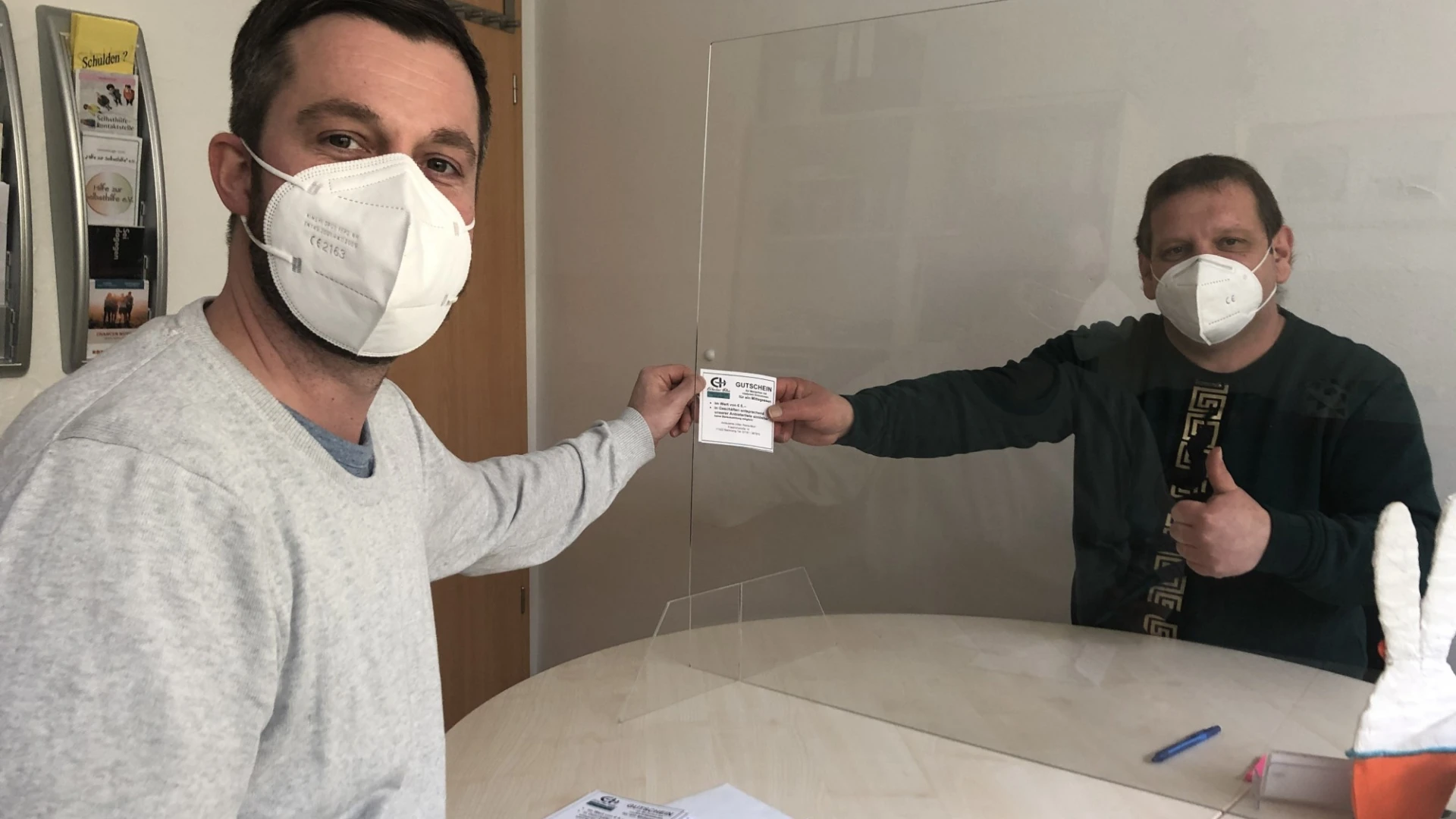 Zwei M&auml;nner mit FFP3-Masken sitzen an einem runden Tisch durch eine durchsichtige Trennwand getrennt. Beide halten ein St&uuml;ck Papier in der Hand und blicken in die Kamera.