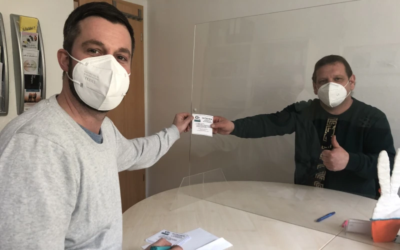 Zwei M&auml;nner mit FFP3-Masken sitzen an einem runden Tisch durch eine durchsichtige Trennwand getrennt. Beide halten ein St&uuml;ck Papier in der Hand und blicken in die Kamera.