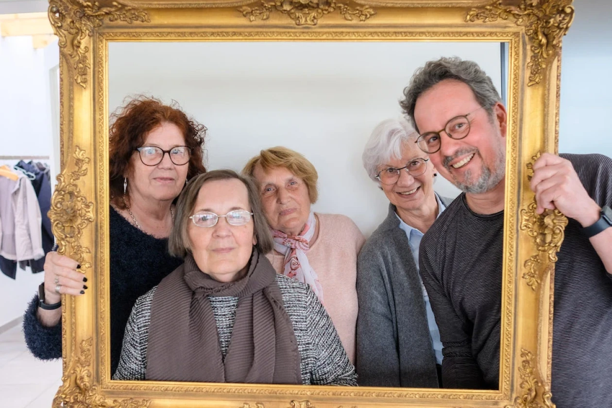 Vier &auml;ltere Frauen und ein Mann mitleren Alters schauen gemeinsam durch einen goldenen Bilderrahmen und l&auml;cheln.