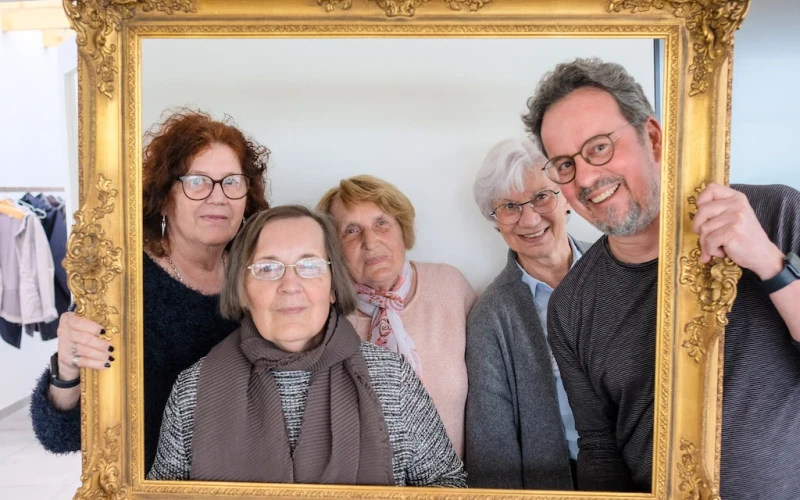 Vier &auml;ltere Frauen und ein Mann mitleren Alters schauen gemeinsam durch einen goldenen Bilderrahmen und l&auml;cheln.