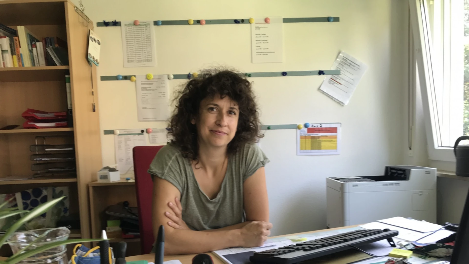 Eine junge Frau mit dunklen lockigen Haaren sitzt hinter einem Schreibtisch voller B&uuml;romaterialien. Im Hintergrund befindet sich ein Schrank, ein Drucker und Dokumente an der Wand.