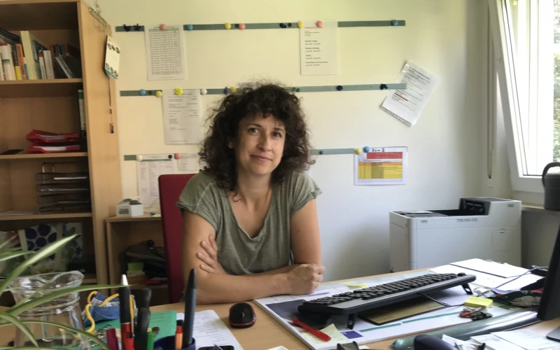 Eine junge Frau mit dunklen lockigen Haaren sitzt hinter einem Schreibtisch voller B&uuml;romaterialien. Im Hintergrund befindet sich ein Schrank, ein Drucker und Dokumente an der Wand.