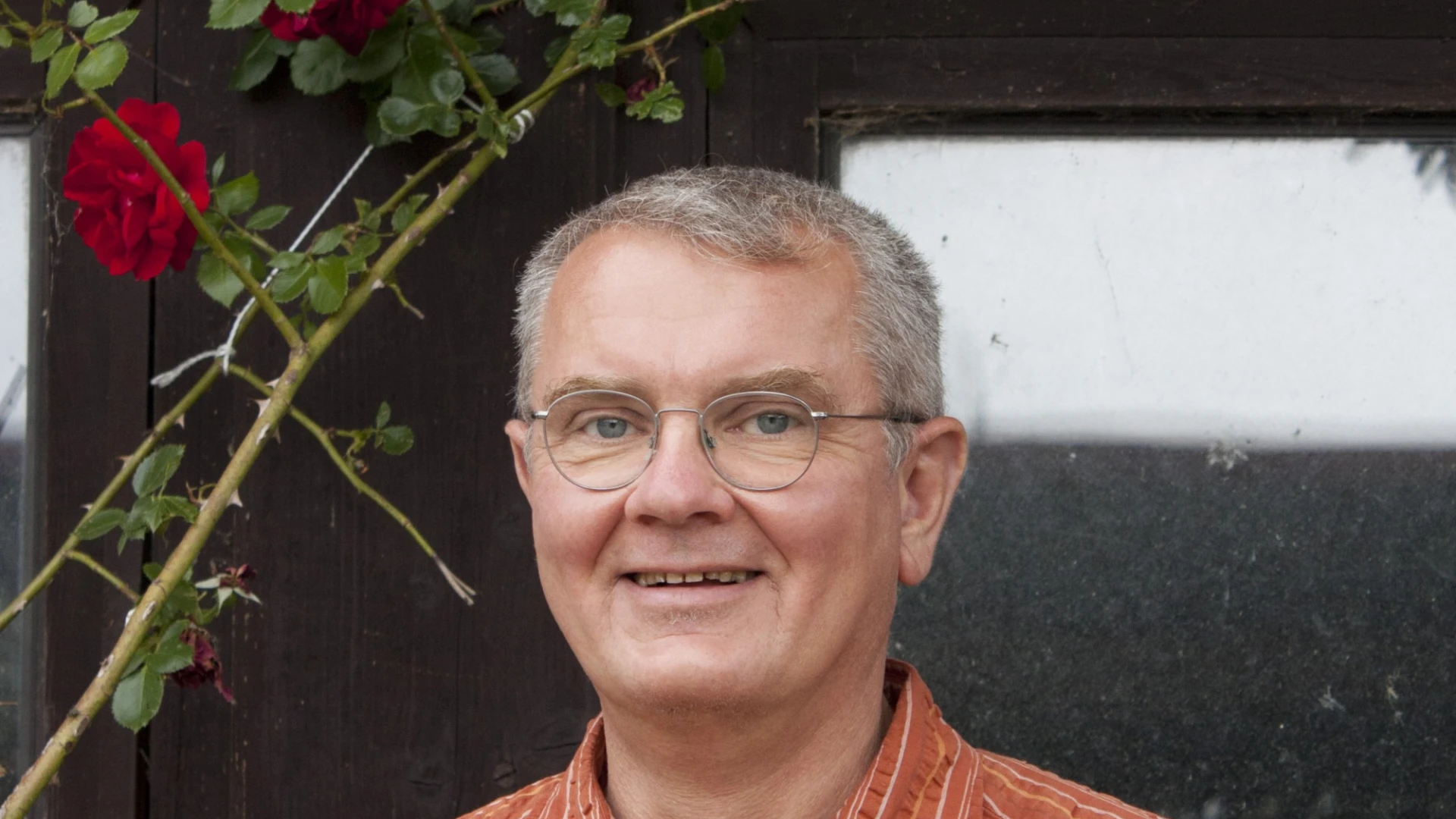 Ein Mann mit Brille und kurzen grauen Haaren, gekleidet mit einem orangefarbenen Hemd l&auml;chelt in die Kamera.