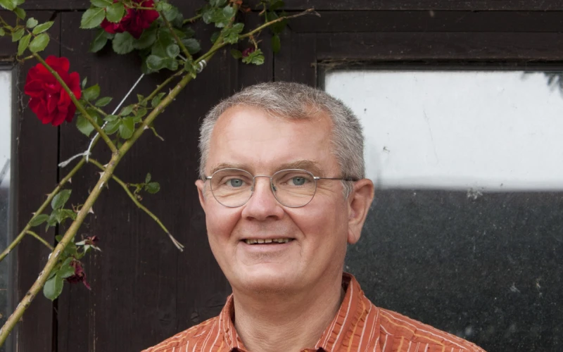 Ein Mann mit Brille und kurzen grauen Haaren, gekleidet mit einem orangefarbenen Hemd l&auml;chelt in die Kamera.