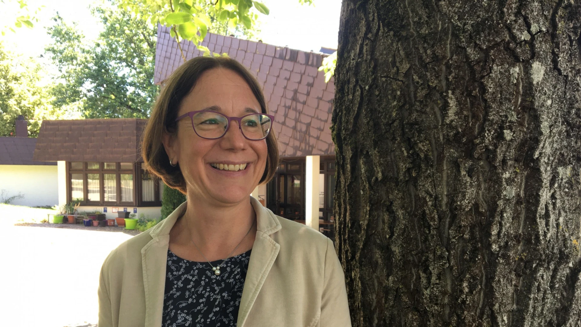 Eine Frau mit Brille und beigem Mantel steht l&auml;chelnd neben einem Baum. Im Hintergrund ist der Eingangsbereich eines Hauses mit Pflanzen zu sehen.