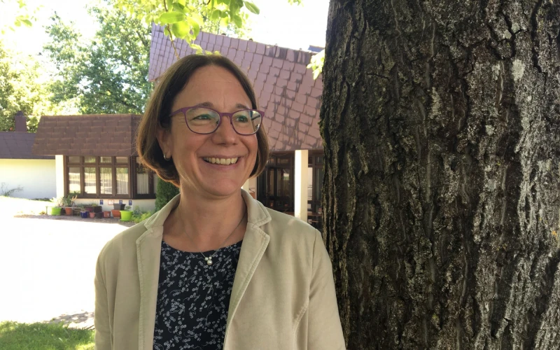 Eine Frau mit Brille und beigem Mantel steht l&auml;chelnd neben einem Baum. Im Hintergrund ist der Eingangsbereich eines Hauses mit Pflanzen zu sehen.