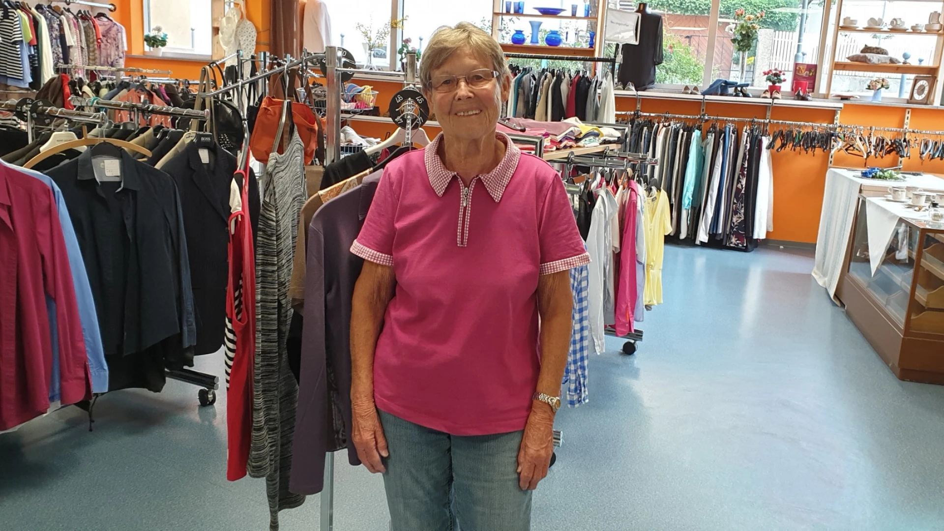 Eine Frau in rosafarbenem Oberteil und einer Brille blickt in die Kamera. Im Hintergrund ist ein Ladengesch&auml;ft zu sehen mit einer Verkaufstheke und mehreren Kleiderst&auml;ndern mit Kleidung best&uuml;ckt.