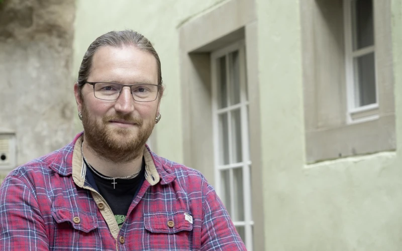 Ein Mann in kariertem Hemd und Brille sitzt auf einem Stein neben einem Geb&auml;ude und l&auml;chelt in die Kamera.