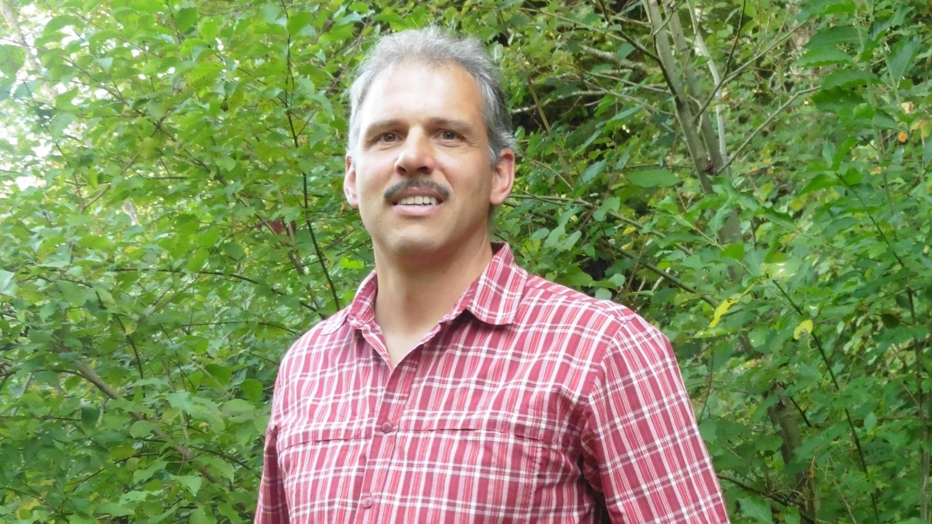 Ein Mann mit Schnauzer, kurzen grauen Haaren und rot kariertem Hemd steht vor einem gr&uuml;nen Hecke.