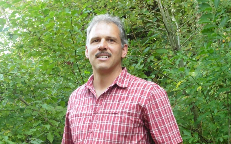 Ein Mann mit Schnauzer, kurzen grauen Haaren und rot kariertem Hemd steht vor einem gr&uuml;nen Hecke.