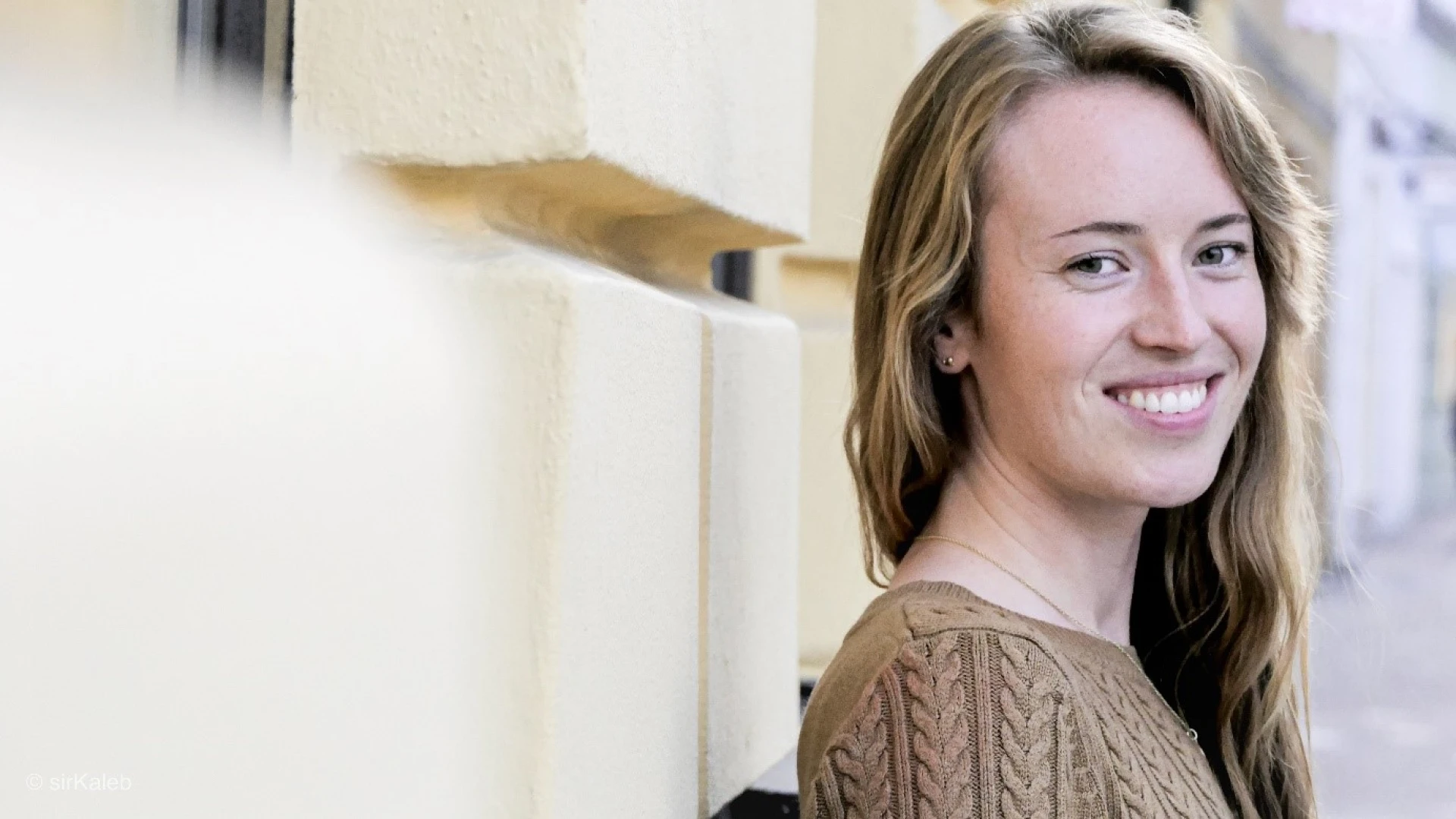 L&auml;chelnde junge Frau mit langen, welligen Haaren, die einen braunen Zopfstrickpullover tr&auml;gt und seitlich in die Kamera blickt.