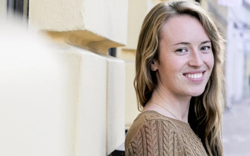 L&auml;chelnde junge Frau mit langen, welligen Haaren, die einen braunen Zopfstrickpullover tr&auml;gt und seitlich in die Kamera blickt.