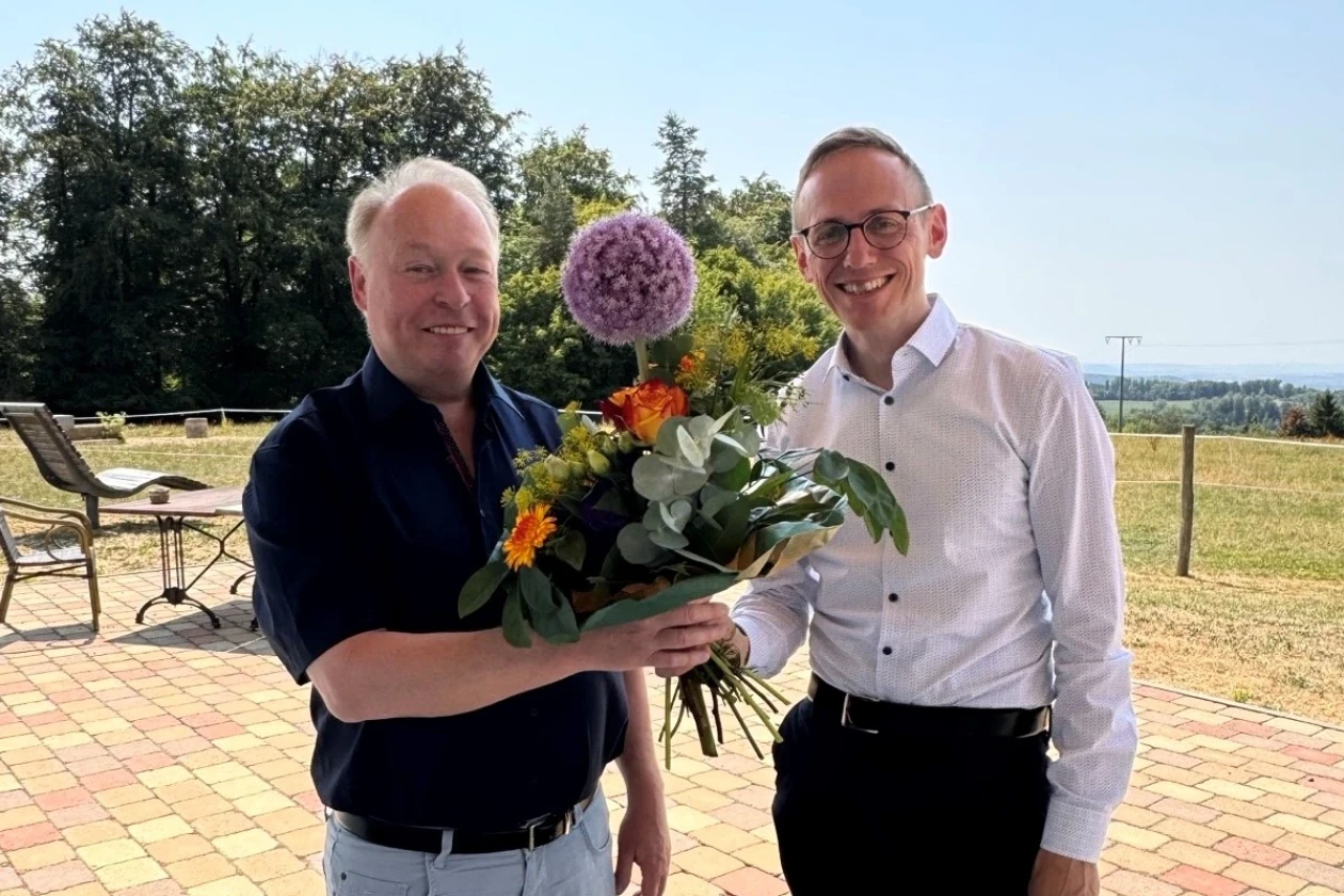 Ein Mann in einem wei&szlig;en Hemd &uuml;berreicht einem anderen Mann mit dunklem Hemd einen Strau&szlig; mit bunten Blumen.