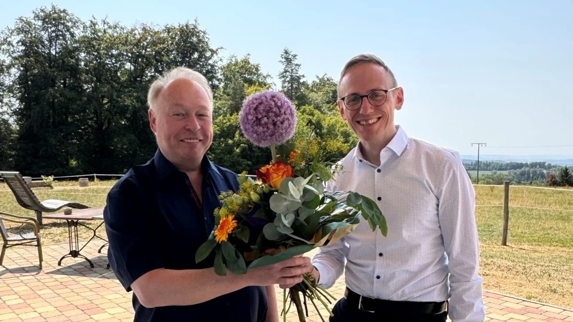 Ein Mann in einem wei&szlig;en Hemd &uuml;berreicht einem anderen Mann mit dunklem Hemd einen Strau&szlig; mit bunten Blumen.