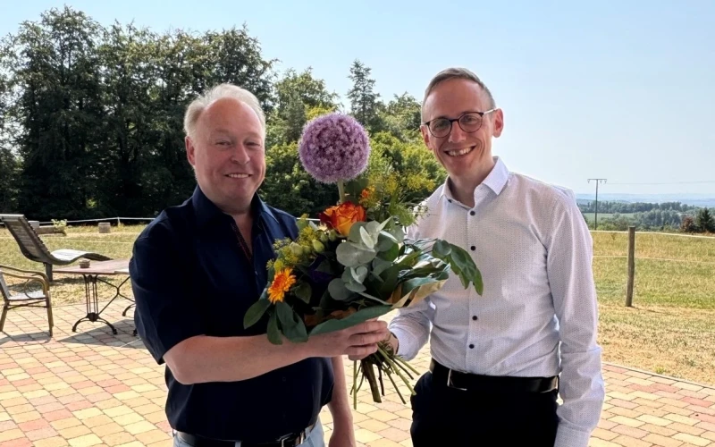 Ein Mann in einem wei&szlig;en Hemd &uuml;berreicht einem anderen Mann mit dunklem Hemd einen Strau&szlig; mit bunten Blumen.