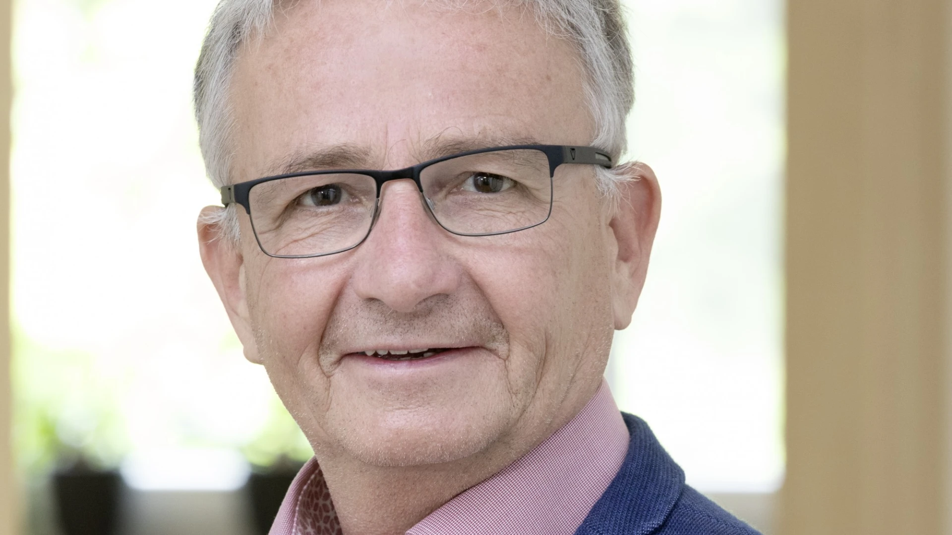 Zu sehen ist ein Mann mit kurzen grauen Haaren, rosa Hemd und einem blauen Jackett. Er tr&auml;gt eine Brille und l&auml;chelt in die Kamera. Im Hintergrund ist ein Fenster zu sehen.