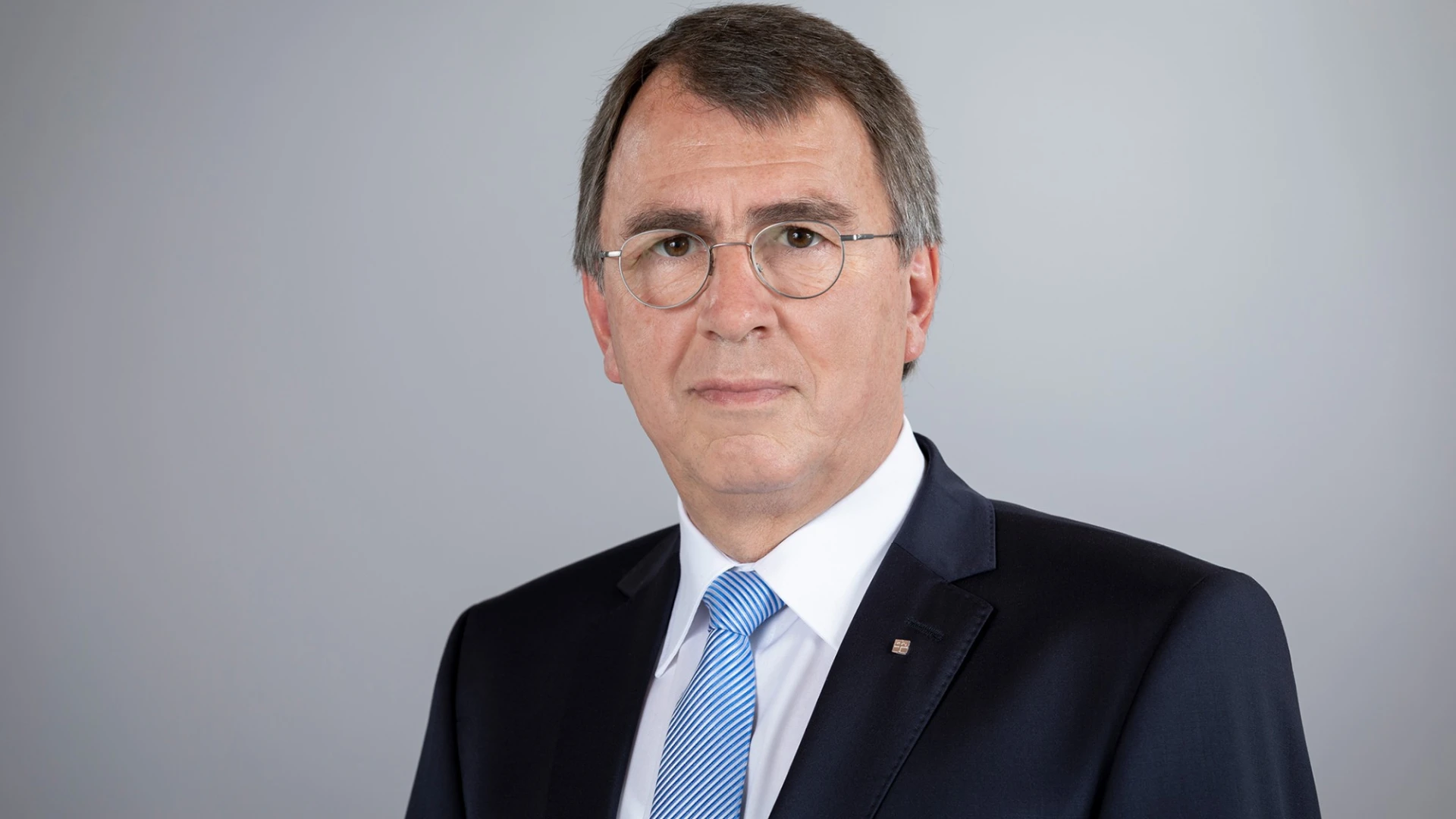 Ein Mann mit Brille, kurzen Haaren, wei&szlig;em Hemd, blau-wei&szlig; gestreifter Krawatte und einem Jackett blickt direkt in die Kamera.