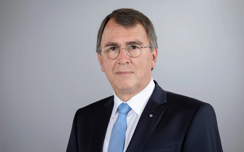 Ein Mann mit Brille, kurzen Haaren, wei&szlig;em Hemd, blau-wei&szlig; gestreifter Krawatte und einem Jackett blickt direkt in die Kamera.