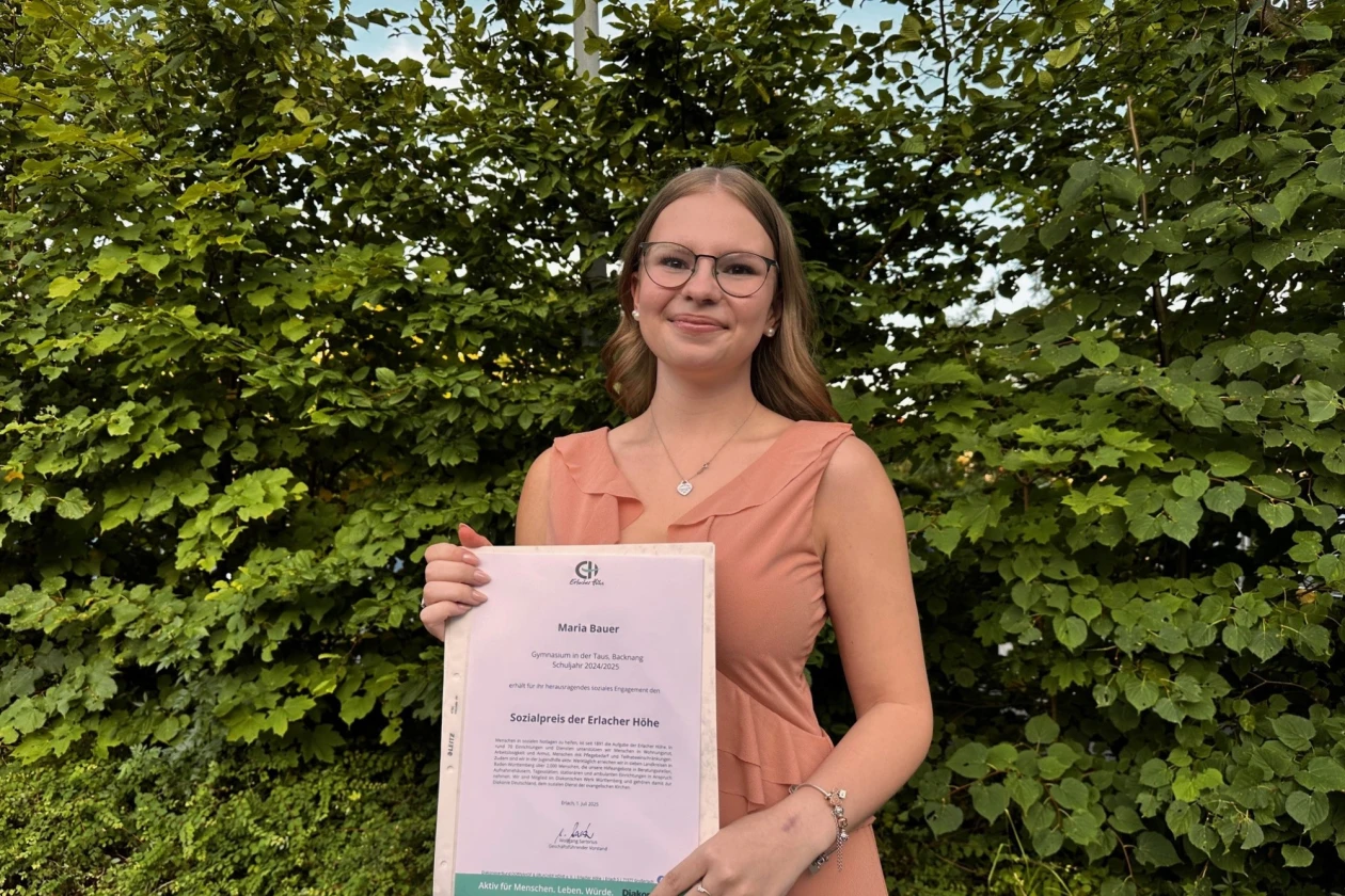 Eine junge Frau mit Brille und langen braunen Haaren steht vor gr&uuml;nem Efeu und h&auml;lt ein Zertifikat in den H&auml;nden