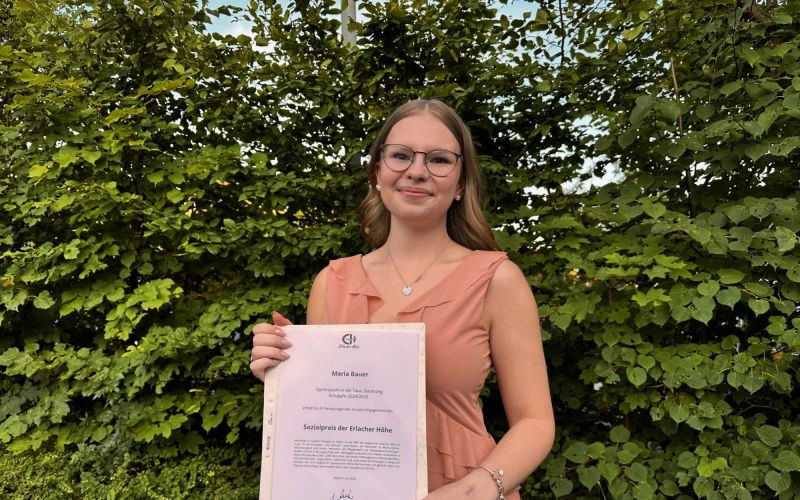 Eine junge Frau mit Brille und langen braunen Haaren steht vor gr&uuml;nem Efeu und h&auml;lt ein Zertifikat in den H&auml;nden