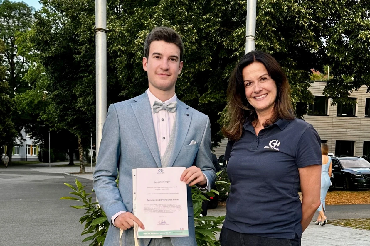 Ein junger Mann in hellem Anzug und Fliege h&auml;lt neben einer Frau in dunklem Poloshirt eine Urkunde mit Zweigen in den H&auml;nden