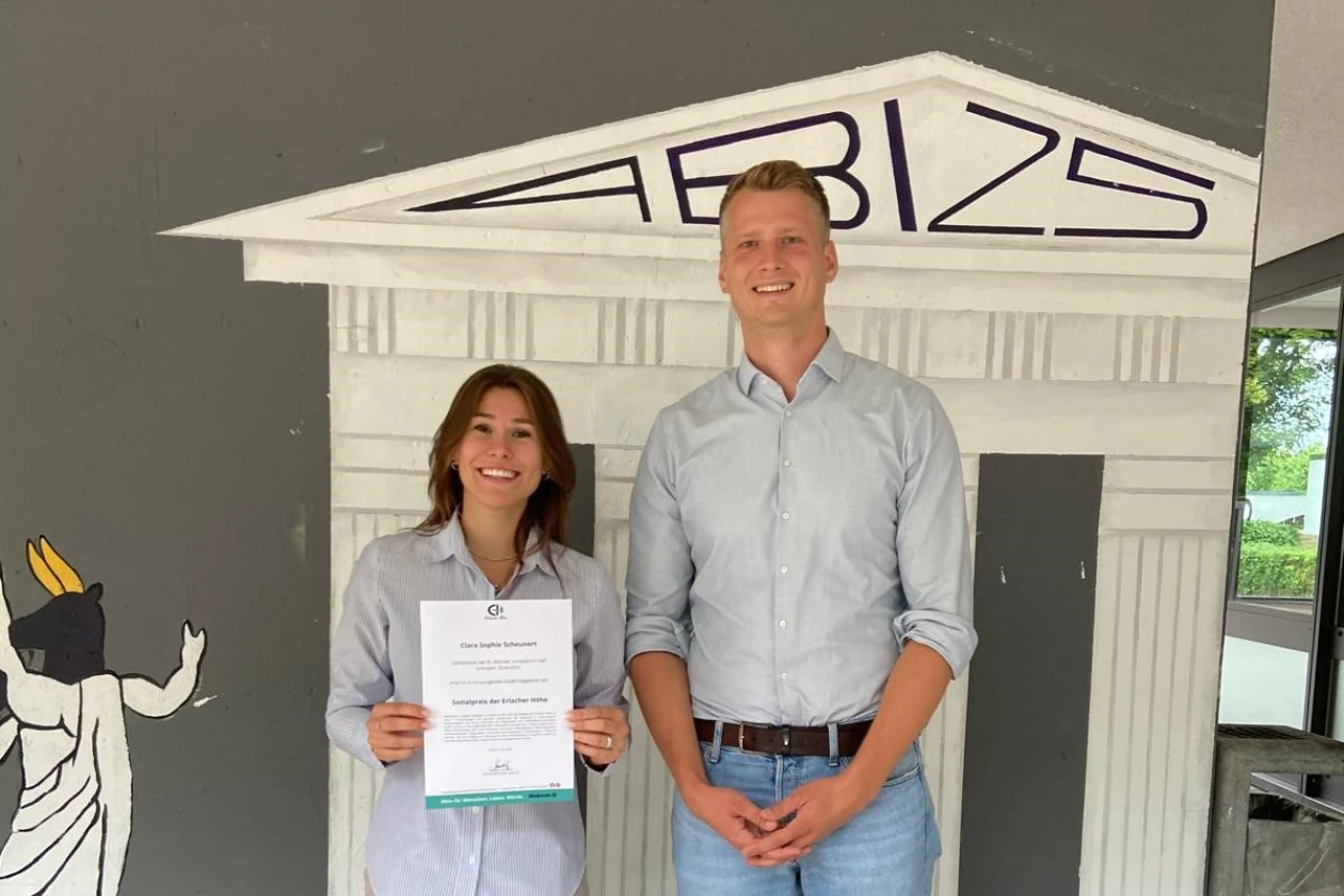 Eine Frau und ein Mann stehen vor einem gro&szlig;en wei&szlig;en Modellhaus und halten gemeinsam ein Zertifikat in die Kamera