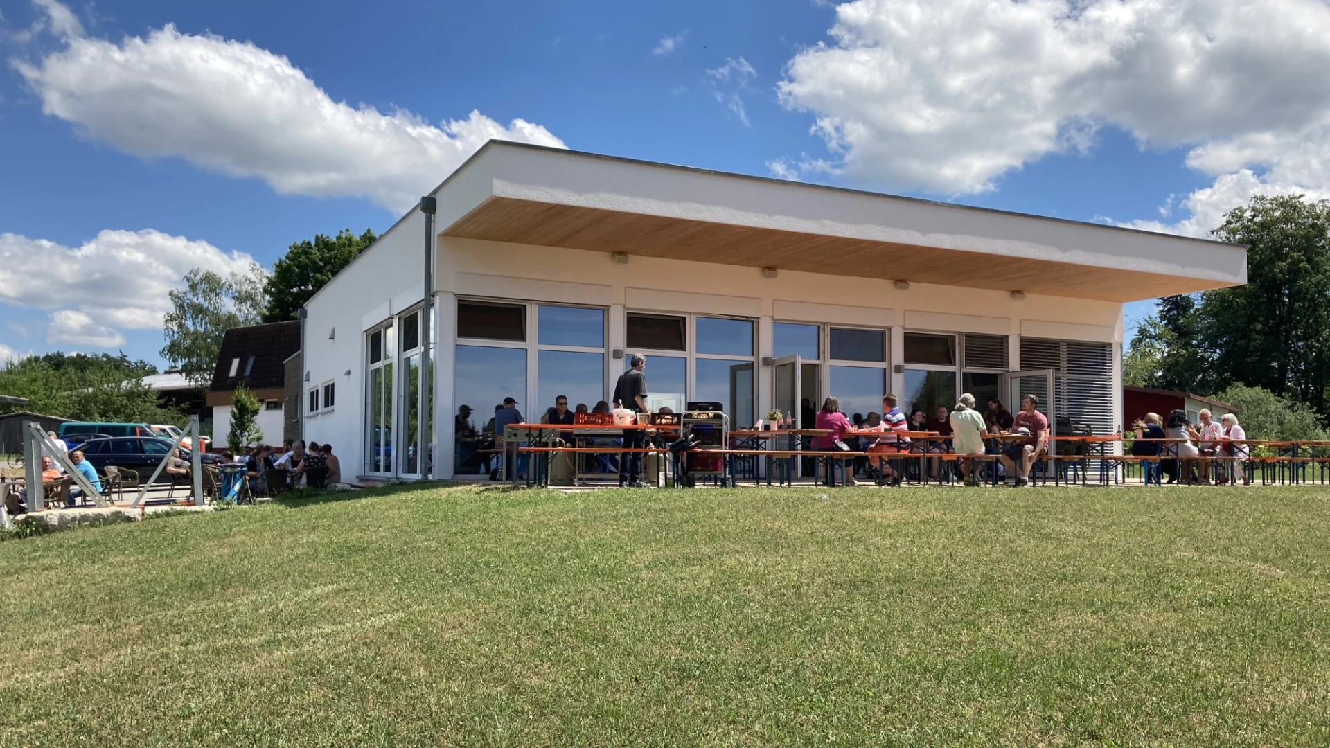 Ein modernes Geb&auml;ude im Gr&uuml;nen bei blauem Himmel mit wei&szlig;en Wolken. Am Geb&auml;ude sind Festzeltgarnituren aufgestellt, an denen Menschen sitzen.