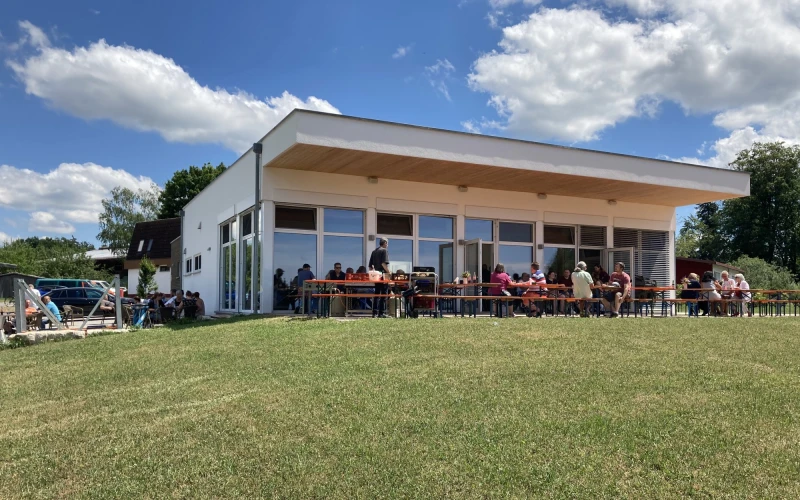 Ein modernes Geb&auml;ude im Gr&uuml;nen bei blauem Himmel mit wei&szlig;en Wolken. Am Geb&auml;ude sind Festzeltgarnituren aufgestellt, an denen Menschen sitzen.