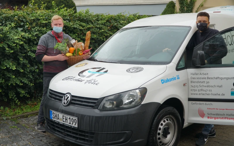 Ein Mann mit Mund-Nasenschutz steht links neben einem wei&szlig;en Transporter und h&auml;lt einen Korb mit Lebensmitteln &ndash; darunter Brot, Obst und Gem&uuml;se &ndash; in der Hand. Ein weiterer Mann steht auf der Fahrerseite und h&auml;lt mit der rechten Hand das Lenkrad.