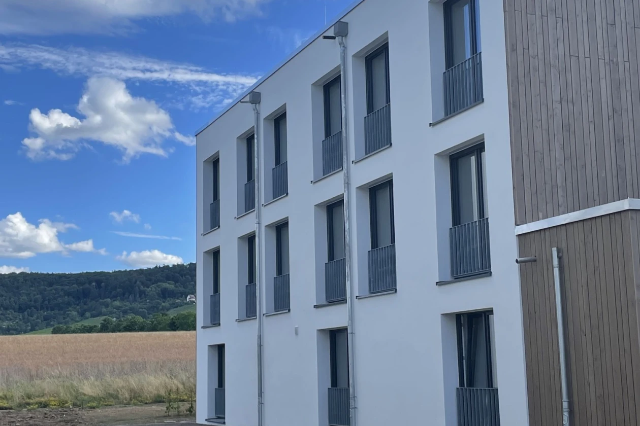 Modernes, dreist&ouml;ckiges Geb&auml;ude mit wei&szlig;er Fassade und Holzanbau, umgeben von einer Baustelle mit Feld und Waldhintergrund. Rechteckige Fenster mit grauen Gel&auml;ndern gliedern die Fassade in ein klares, geometrisches Muster.