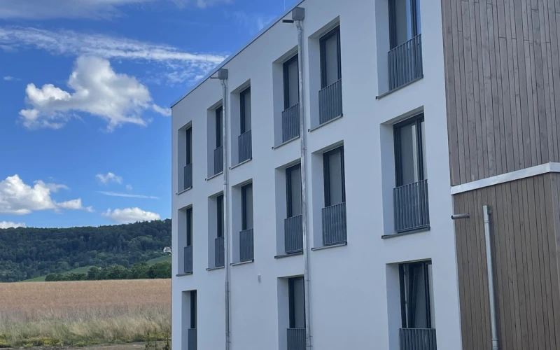 Modernes, dreist&ouml;ckiges Geb&auml;ude mit wei&szlig;er Fassade und Holzanbau, umgeben von einer Baustelle mit Feld und Waldhintergrund. Rechteckige Fenster mit grauen Gel&auml;ndern gliedern die Fassade in ein klares, geometrisches Muster.