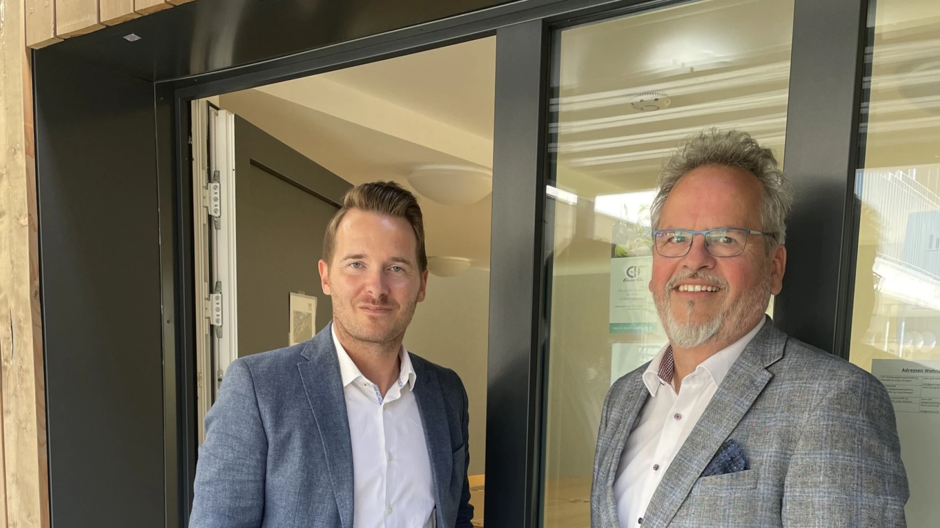 Zwei M&auml;nner am Eingang eines Geb&auml;udes. Beide tragen Jackett und Hemd und l&auml;cheln in die Kamera.