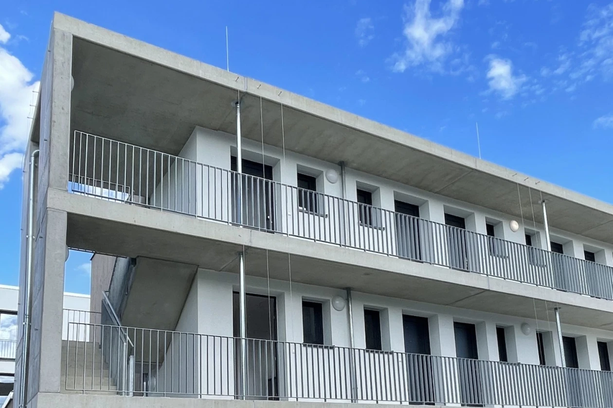 Dreist&ouml;ckiges, modernes Betongeb&auml;ude mit wei&szlig;en Gel&auml;ndern und Au&szlig;entreppen, rechteckigen Fenstern und T&uuml;ren, umgeben von gepflastertem Boden und einer kleinen Rasenfl&auml;che vor blauem Himmel.